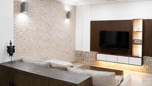 A modern living room with a large L-shaped sectional sofa, a wooden cabinet, a flat-screen TV mounted on a wooden wall, recessed ceiling lights, and a ceiling fan. - Luxor Garden Residence (Accra)