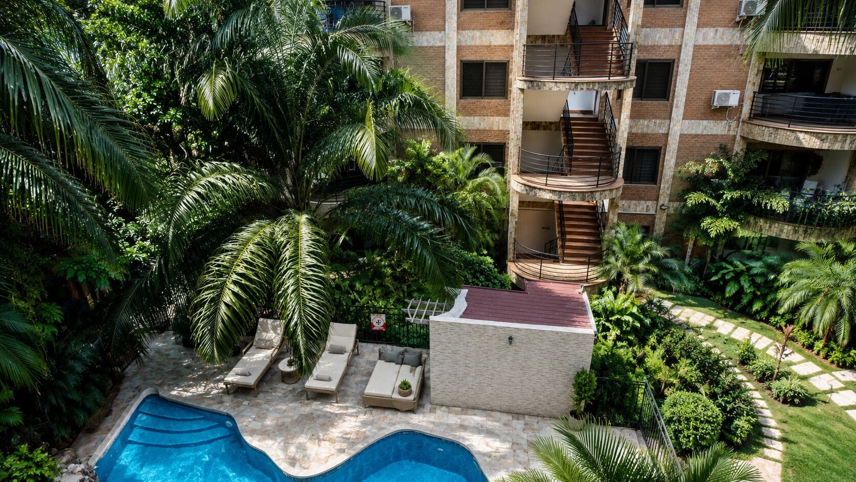 A rooftop pool with lounge chairs, surrounded by lush greenery and a multi-story building with balconies.