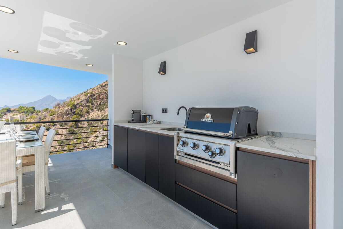 An outdoor kitchen with a grill, dining table, and chairs, overlooking a mountainous landscape.