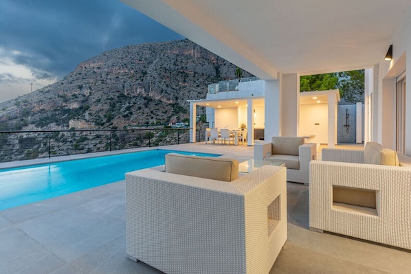 A modern outdoor pool area with white wicker furniture, a view of a mountainous landscape, and a clear sky.