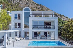 A modern two-story house with a swimming pool, outdoor seating, and a view of a mountainous landscape.