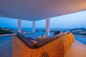 A balcony with a woven sofa, a small round table, and a view of the city at dusk.