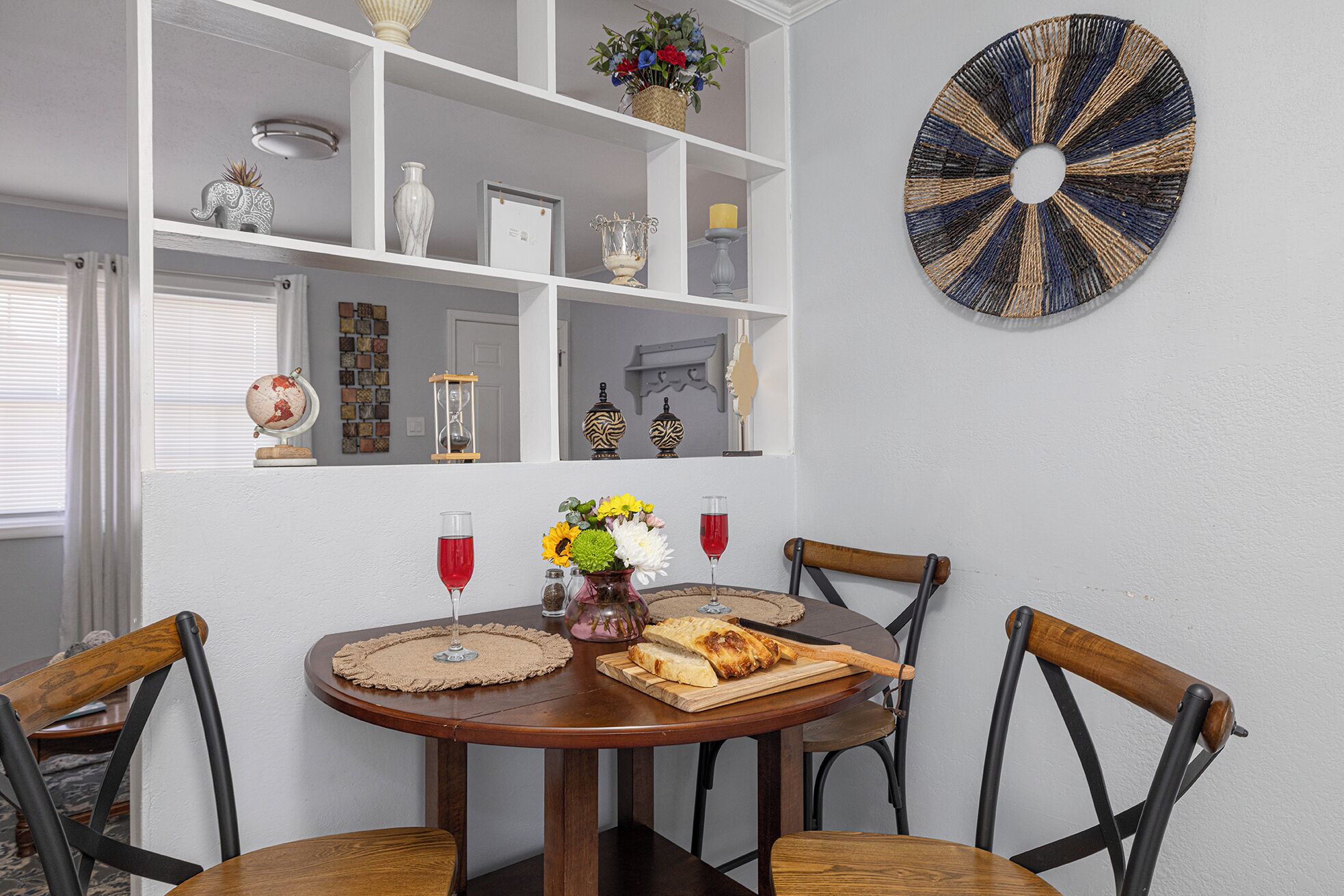 Een eetruimte met een ronde houten tafel, vier houten stoelen, een witte plank met decoratieve voorwerpen en een rond kunstwerk aan de muur.