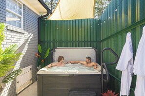 Ein Whirlpool mit zwei Personen, umgeben von einem grünen Zaun und einer weißen Mauer.