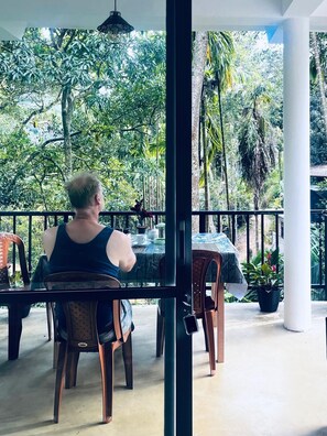 A person seated at a table with a view of dense tropical foliage. - Mithun Homestay (Ella)