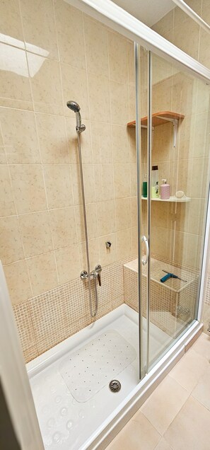 Un espace douche clos par des parois de verre, doté d’un pommeau de douche à main, d’une porte en verre et d’une étagère pour les articles de toilette.