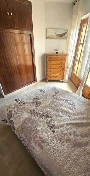 Een slaapkamer met een groot bed, een houten garderobe, een houten dressoir, een schilderij aan de muur en een raam met gordijnen.