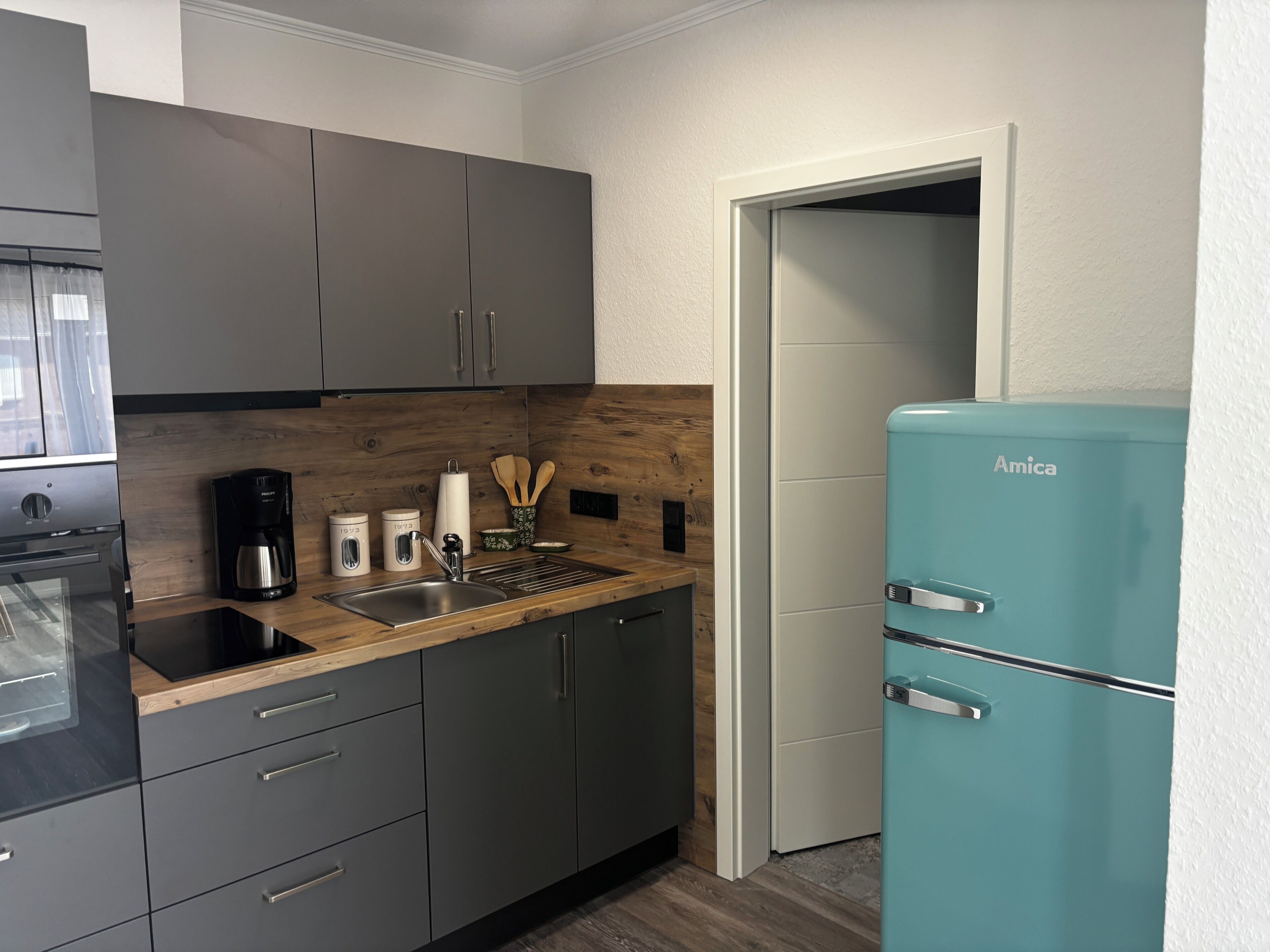 Eine Küche mit grauen Schränken, einer Kaffeemaschine, einem Spülbecken und einem Amica-Kühlschrank in Türkis.