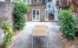 A patio with a wooden table and chairs, a brick wall, and a glass door leading to a balcony. - Sunny, 2bed 2bath, garden apartment near Hyde Park (London)