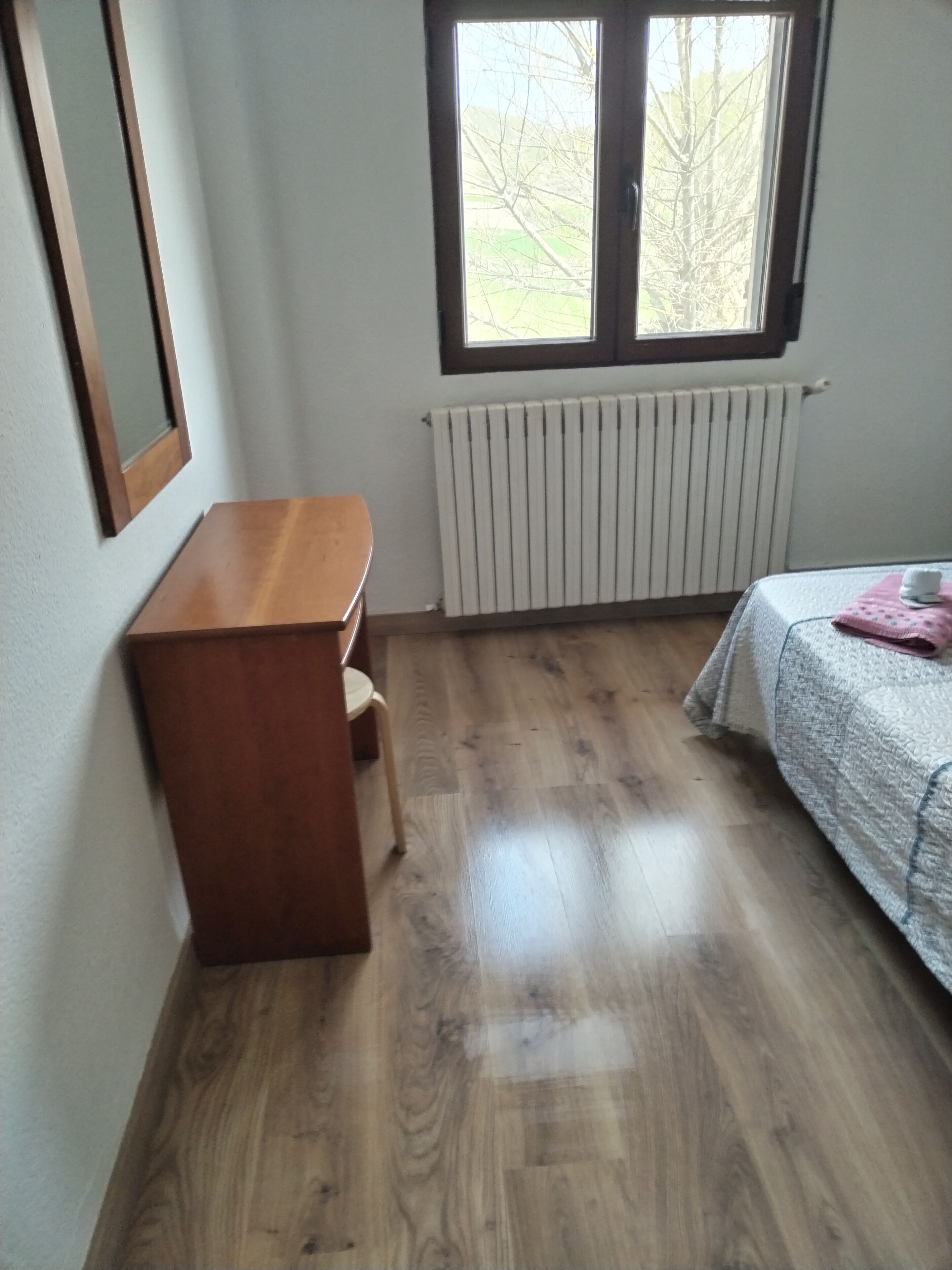 Ein Zimmer mit Schreibtisch aus Holz, Heizkörper, einem Bett mit gesteckter Decke und einem Fenster mit Blick auf Bäume.