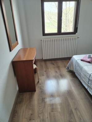 Ein Zimmer mit Schreibtisch aus Holz, Heizkörper, einem Bett mit gesteckter Decke und einem Fenster mit Blick auf Bäume.