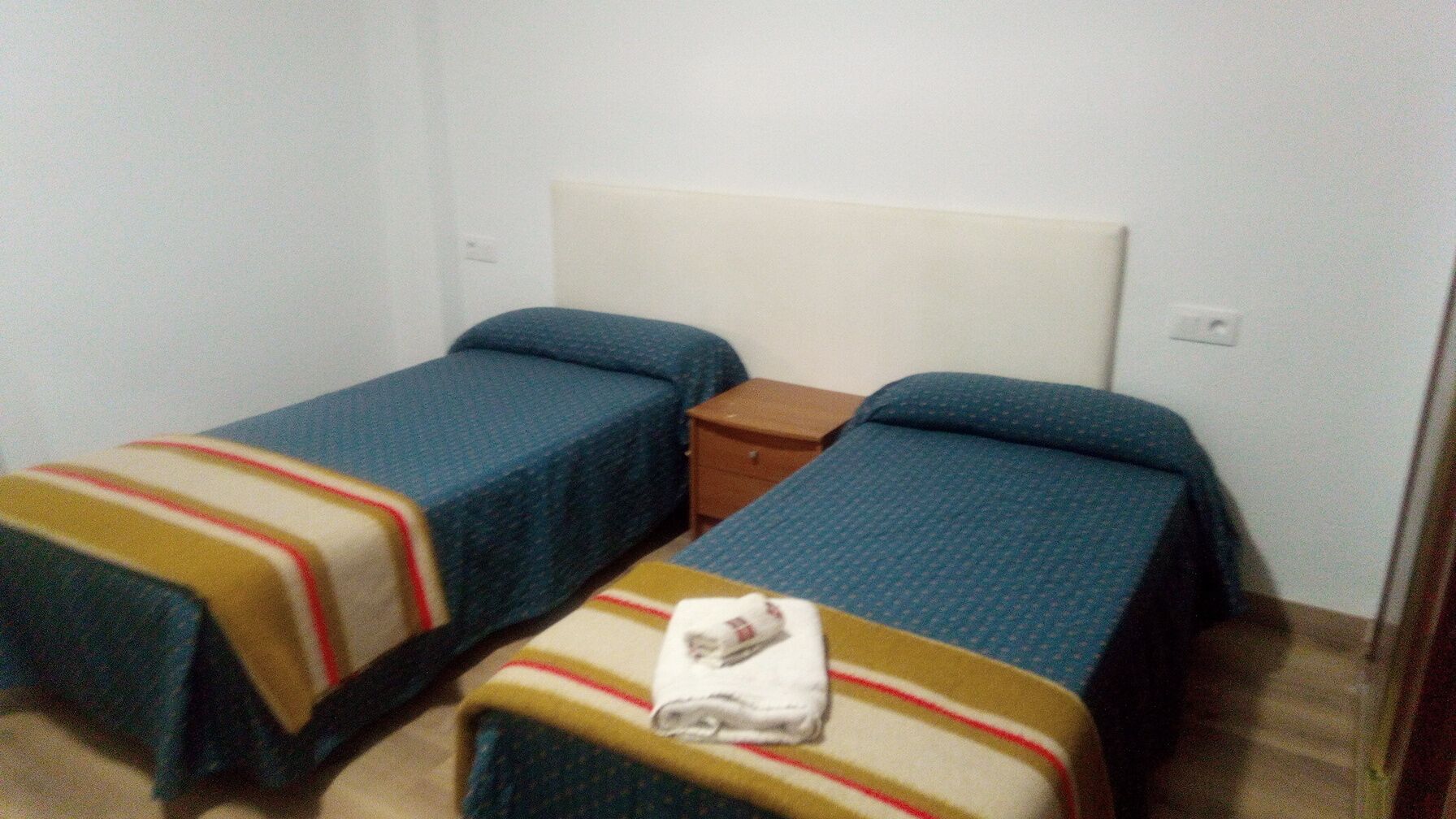 Una habitación con dos camas individuales, una mesita de noche de madera y un cabecero blanco.