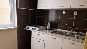 Kompakti keittiö, jossa on kaasuliesi, vedenkeitin ja pesuallas, valkoiset kaapit, tumma laattaseinä ja kuvioitu matto.