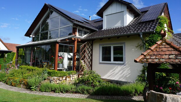 Ein Haus mit einem großen Wintergarten, einem Ziegeldach und einem Garten mit unterschiedlichen Pflanzen und einer Steinmauer.