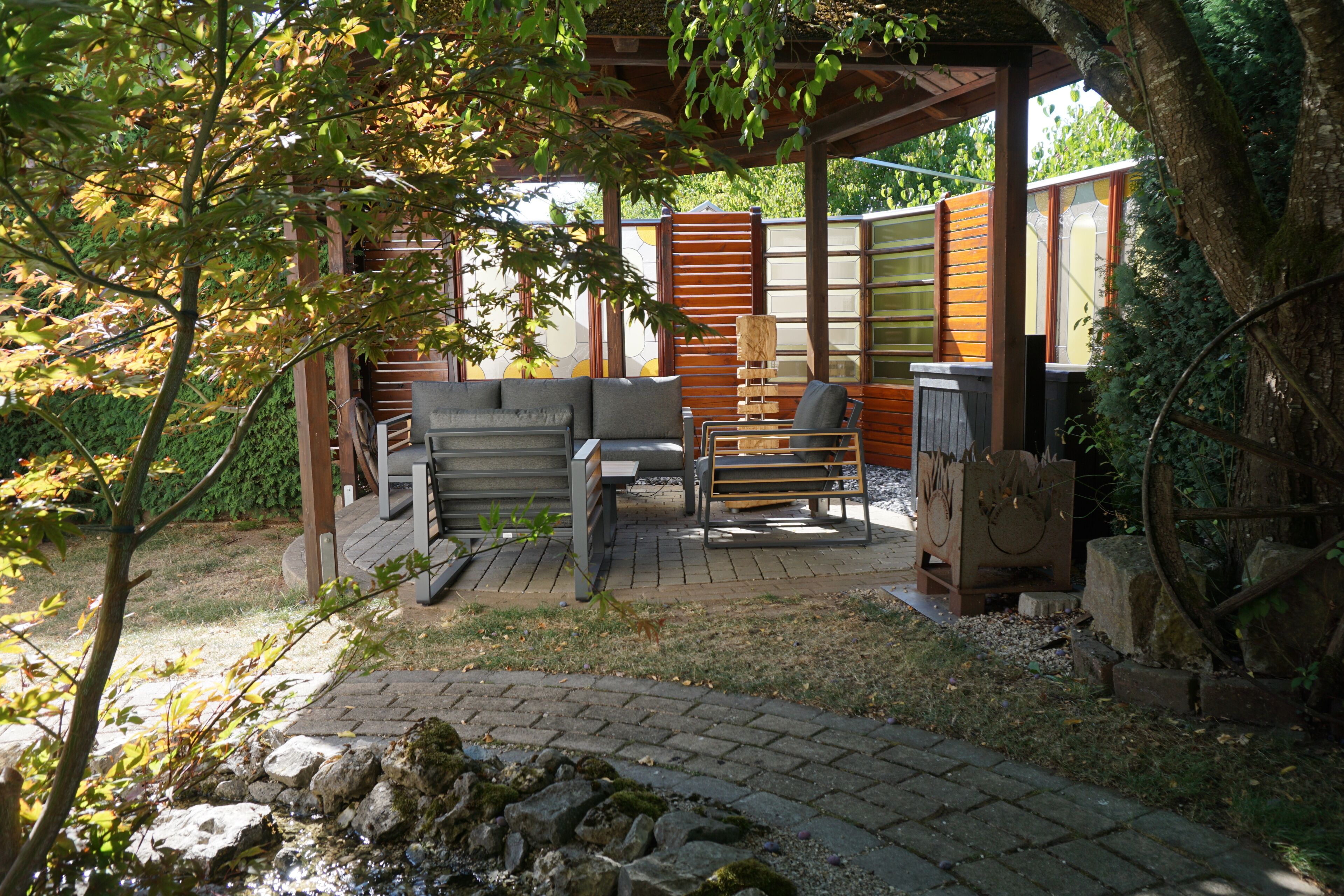 Un espace extérieur aménagé, abrité et doté d’un canapé et de fauteuils, d’une petite table et d’une structure décorative en bois.