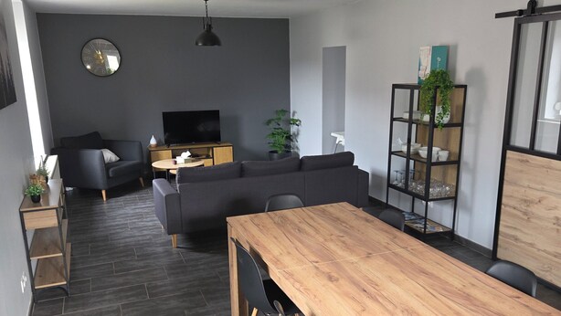Moderni olohuone, jossa on puinen ruokapöytä, mustat tuolit, harmaa sohva, televisio ja viherkasvi.