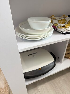 A white shelf with stacked white plates and bowls, a George Foreman grill, and a patterned cloth. - Modern Tiny House in conveniently located Tampa Neighborhood. (Tampa)