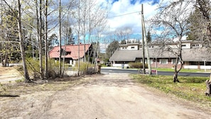 Une route de campagne menant à des maisons aux toits rouges et gris, entourées d’arbres et bordée d’un ciel dégagé.