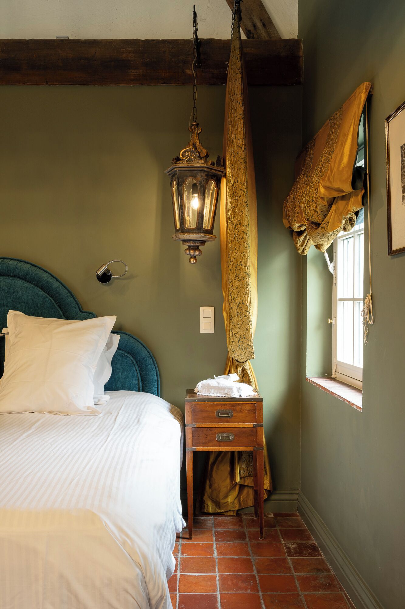 Een slaapkamer met een bed, een nachtkastje, een hanglamp, een raam met gordijnen en een lichtschakelaar aan de muur.