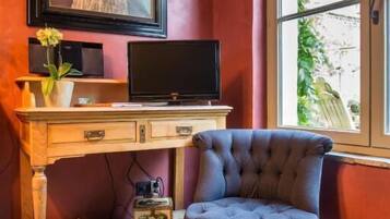 A room with a wooden desk, a television, a chair, a framed picture, and a window with a view of greenery.