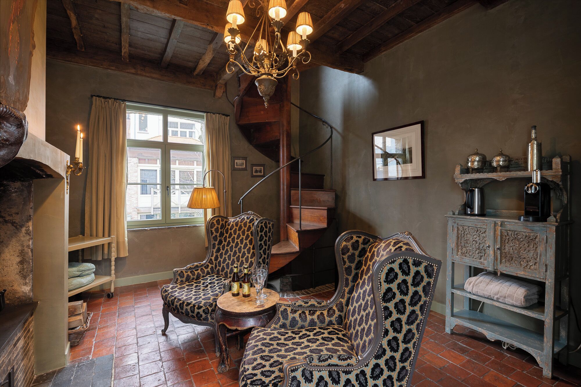 A cozy room with a fireplace, a chandelier, a staircase, and a seating area with patterned upholstery.