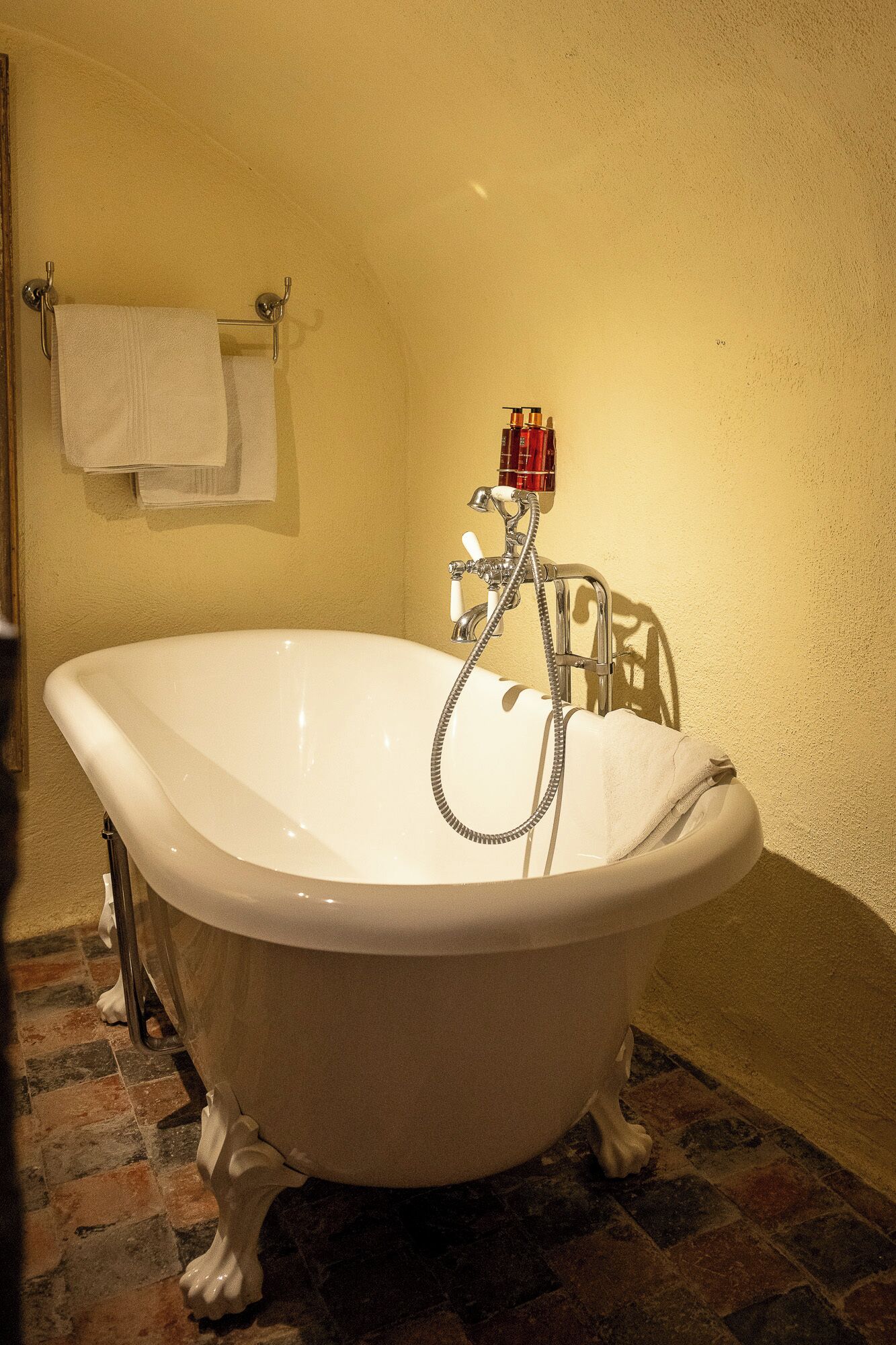 Een ligbad op pootjes met een douchekop, een handdoekhouder met een witte handdoek, en een fles shampoo.