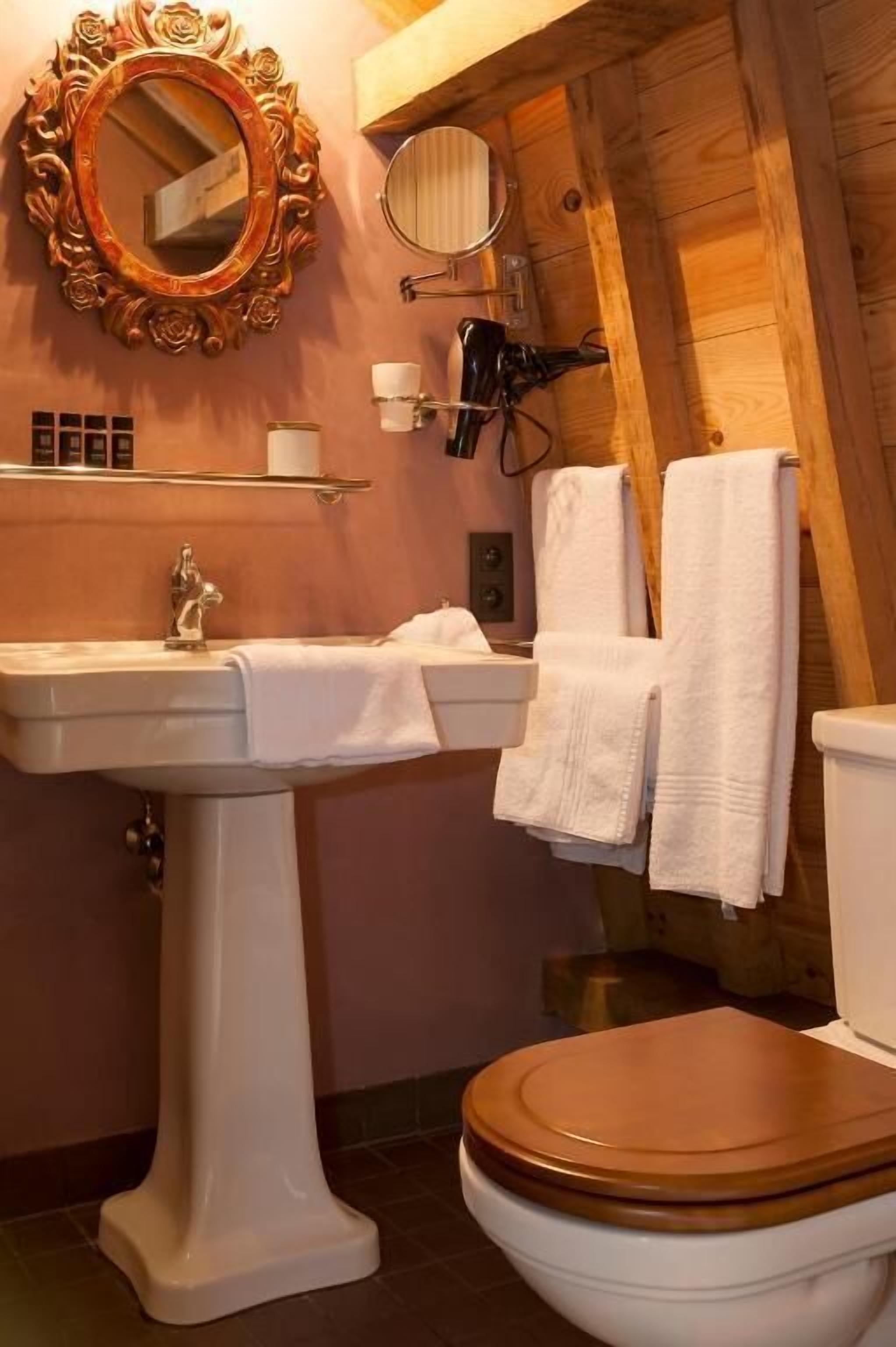 Un bagno con un lavandino su piedistallo, un water con sedile in legno, uno specchio con cornice decorata e un portasciugamani.