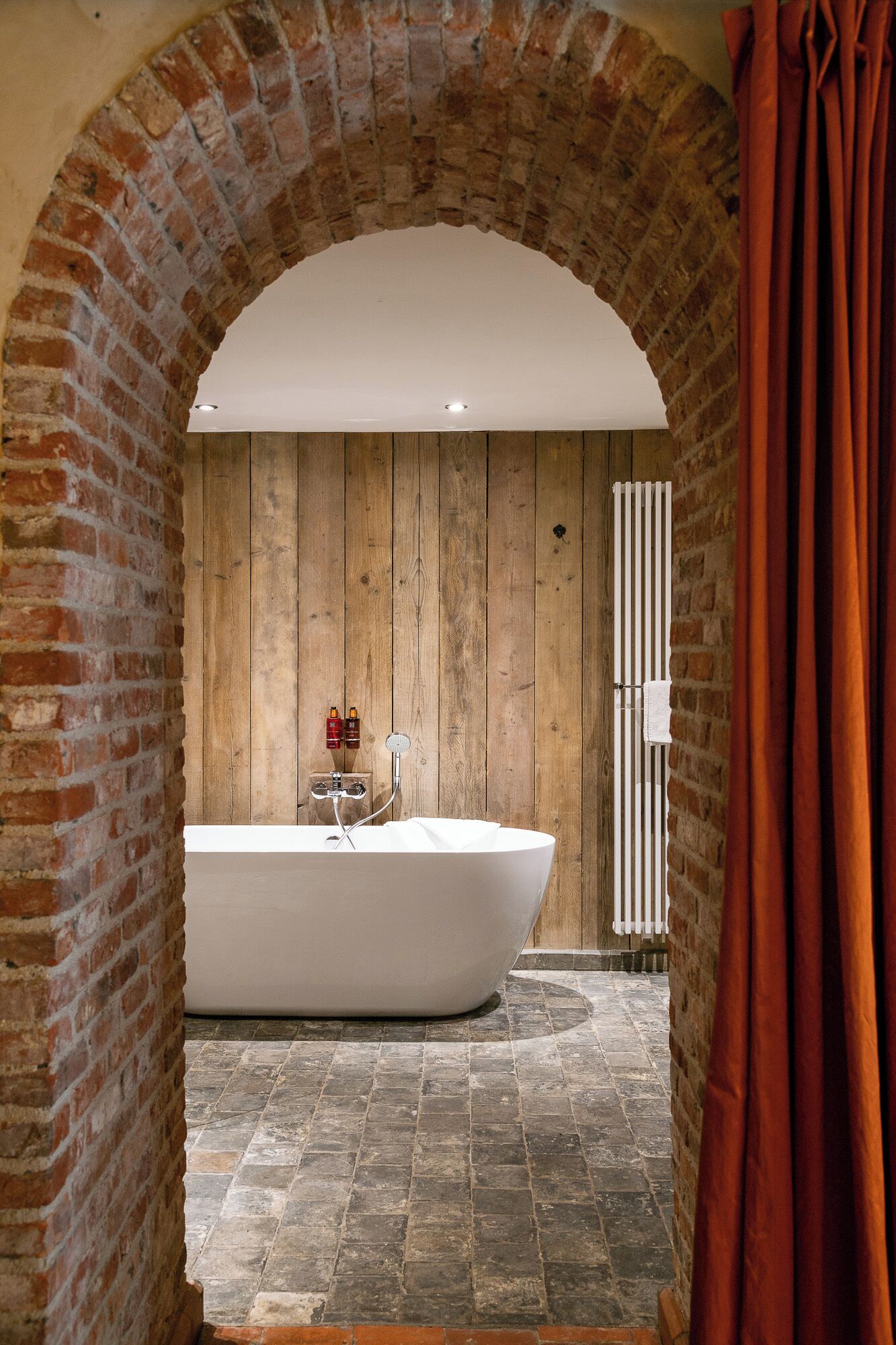 A bathroom with a freestanding bathtub, wooden panel walls, and a brick archway.