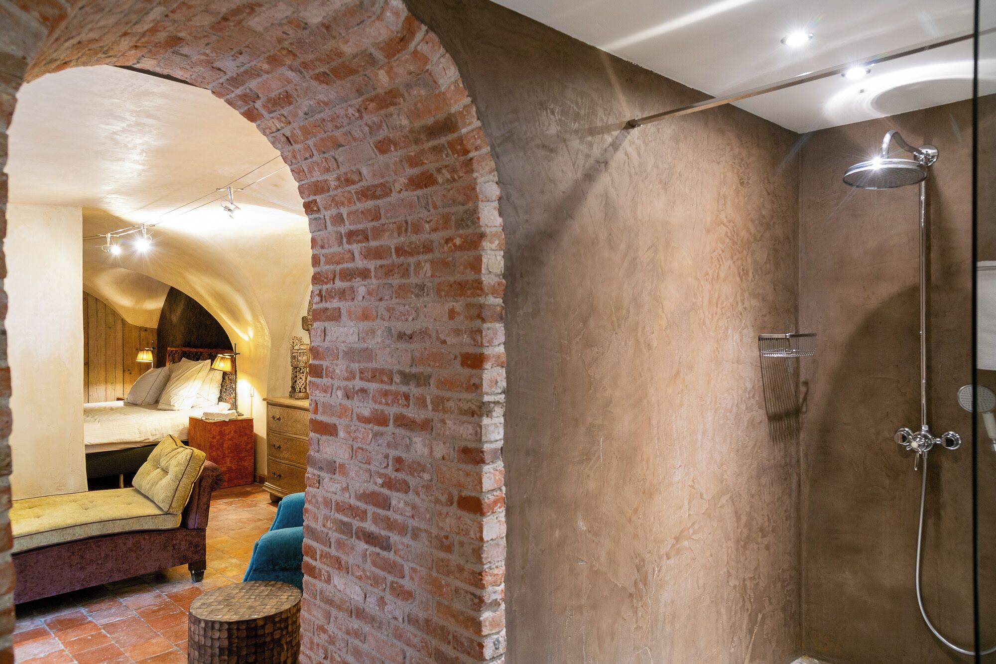 A room with a brick wall, a bed, a sofa, a small table, and a shower area.