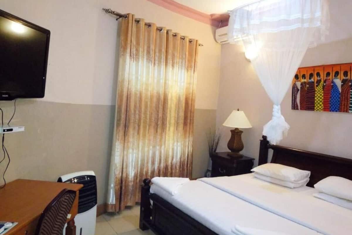 Habitación de hotel con cama, escritorio, televisor, cortina, mesita de noche y aire acondicionado instalado en la pared.