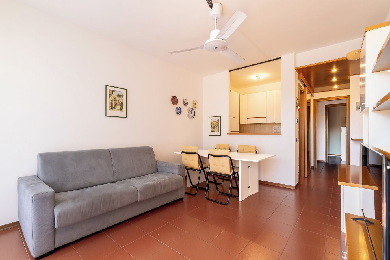Un soggiorno con un divano grigio, un tavolo da pranzo con sedie, una ventola a soffitto e un angolo cottura con pensili.