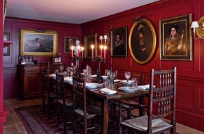 A formal dining room with a long table set for six, surrounded by chairs, with framed portraits on the walls and a red paneled background.