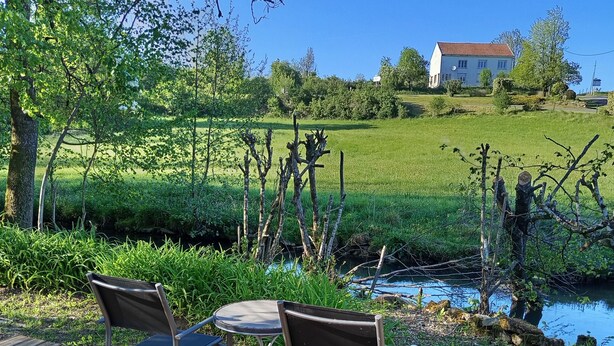 A wooden deck with two chairs and a table, a small stream, and a house in the background. - Maisonnette le poulailler (Vaux-Villaine)
