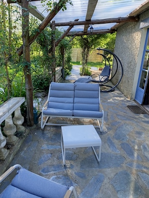 Eine Steinterrasse mit einer grauen Couch, einem weißen Hocker und einem Gartenbereich mit einer Holzw pergola und einem Metallstuhl.