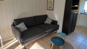 A living room with a sofa, two side tables, a framed newspaper on the wall, and a kitchen area with cabinets. - Maisonnette le poulailler (Vaux-Villaine)
