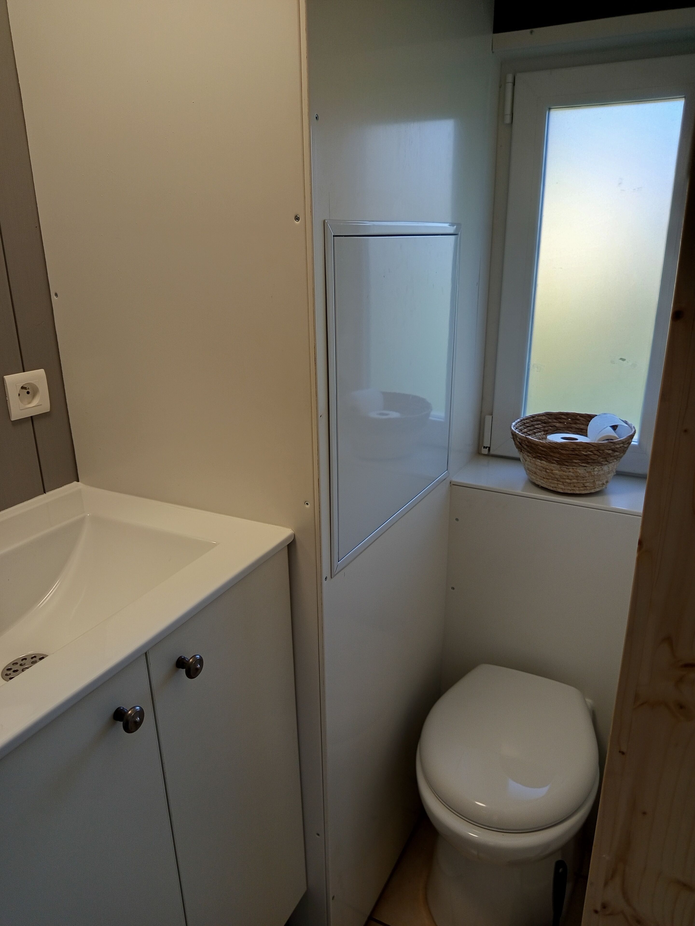 A compact bathroom with a white sink, a toilet, and a window with a basket on the sill.
