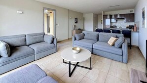 Ein modernes Wohnzimmer mit zwei grauen Sofas, einem runden Couchtisch und einer Küche im Hintergrund.
