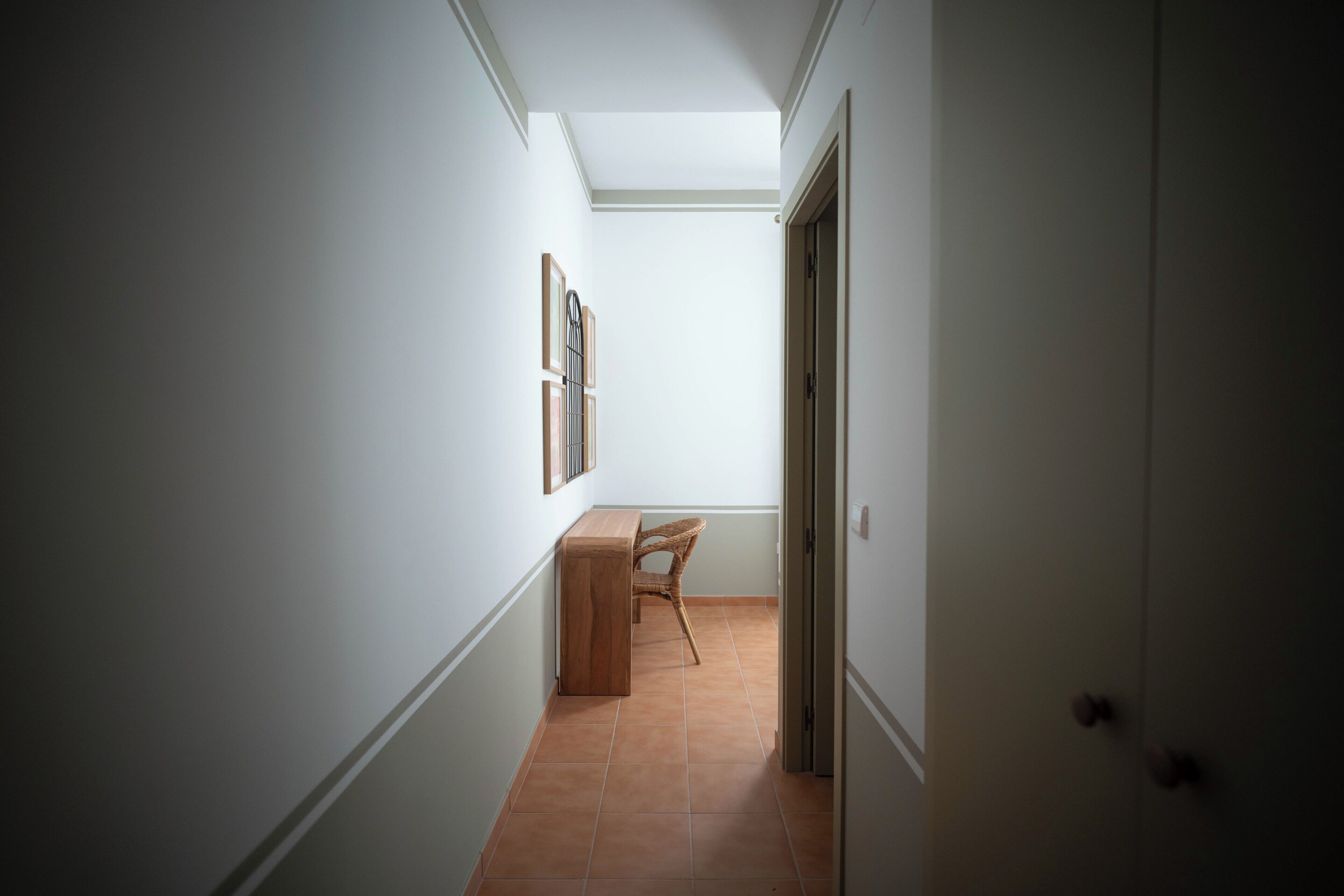 Un pasillo con una puerta a la derecha, una silla y un estante en la pared con objetos enmarcados.