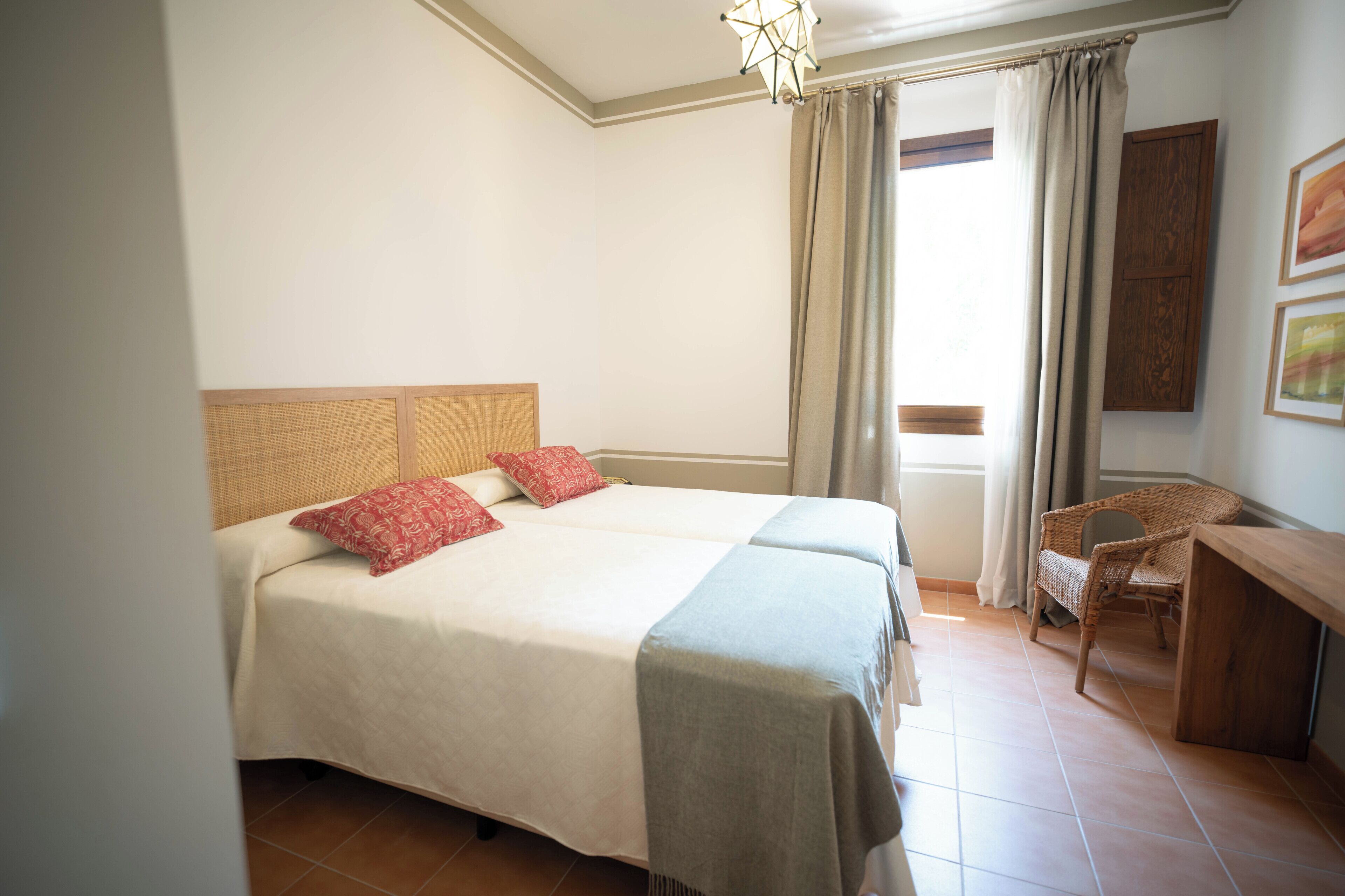 Una cama doble con cabecera, dos almohadas, una mesita de noche, una silla y una ventana con cortinas.