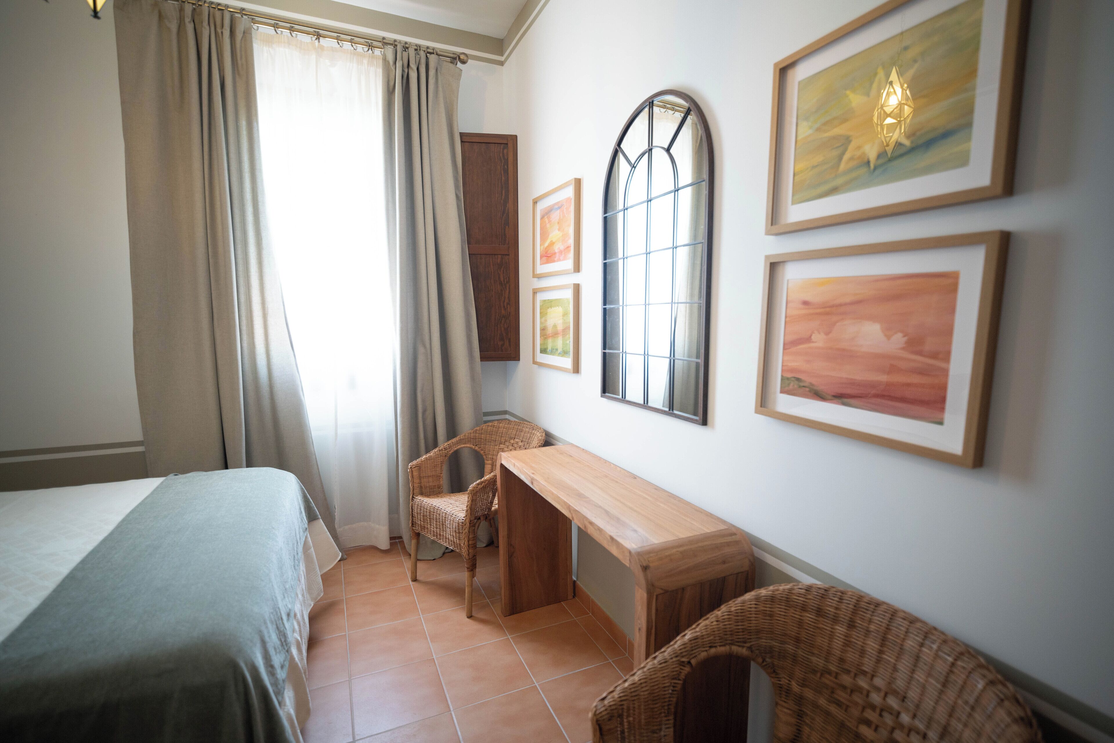 Un dormitorio con cama, escritorio, dos sillas, ventana con cortinas y cuadros en la pared.