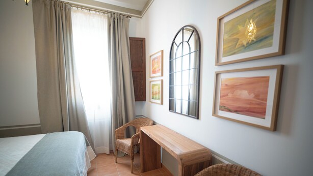 Un dormitorio con cama, escritorio, dos sillas, ventana con cortinas y cuadros en la pared.