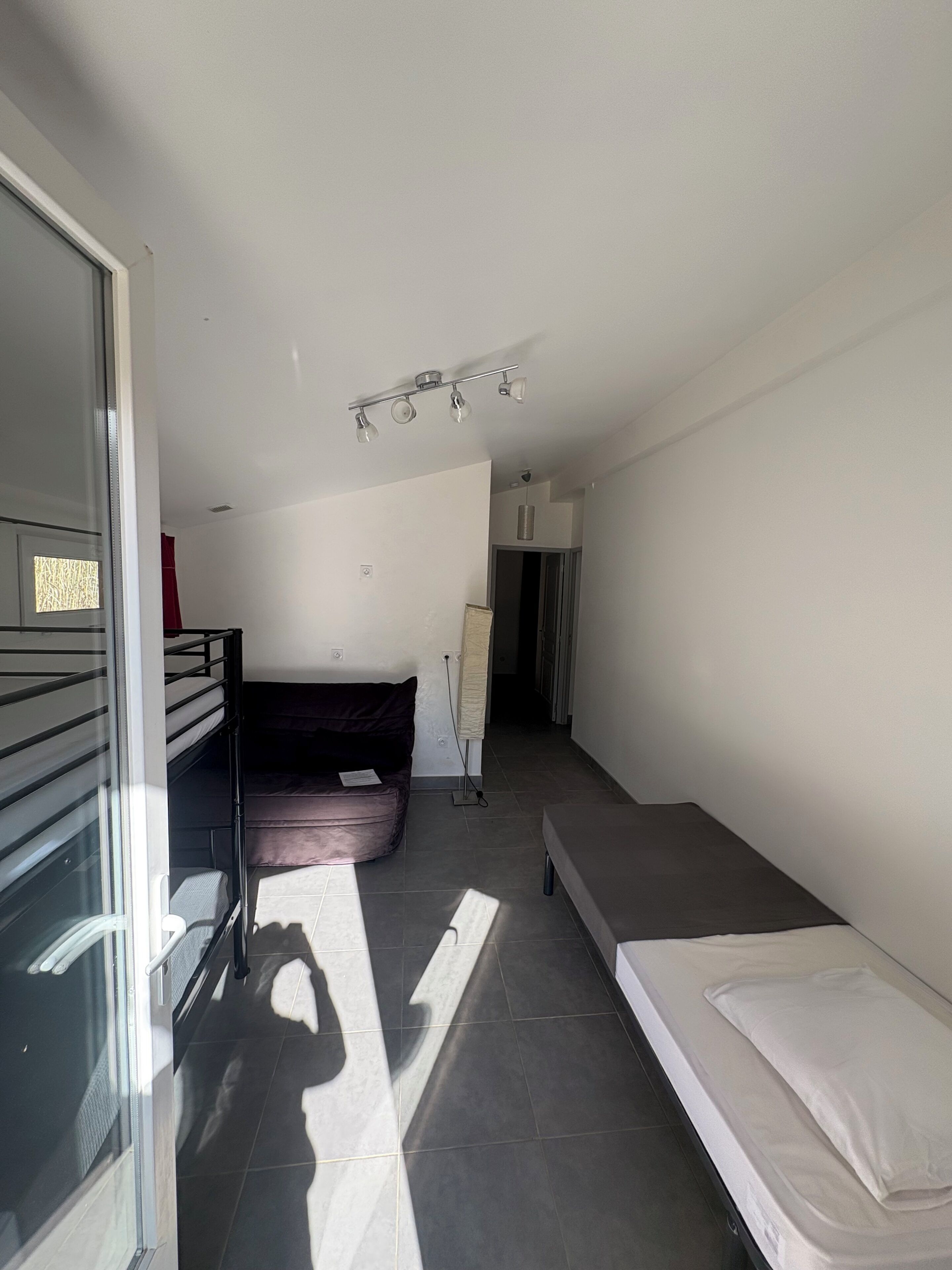 A modern, minimalistic room with a bed, a sofa, a small table, and a glass door leading to an outdoor area.
