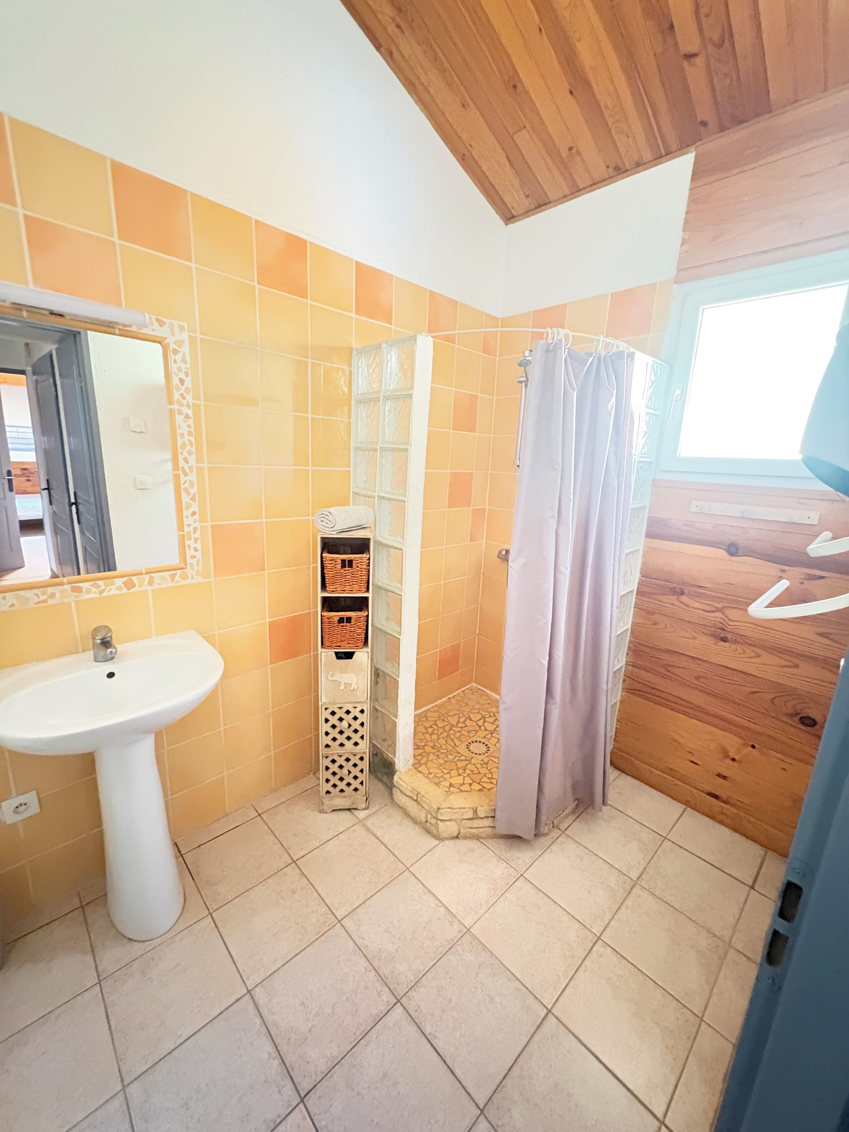 Un baño con techo de madera, un lavamanos blanco, una ducha con puerta de madera y una ventana.