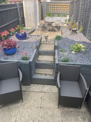 En terrasse med udendørs møbler, en lille hund og en have med forskellige planter.
