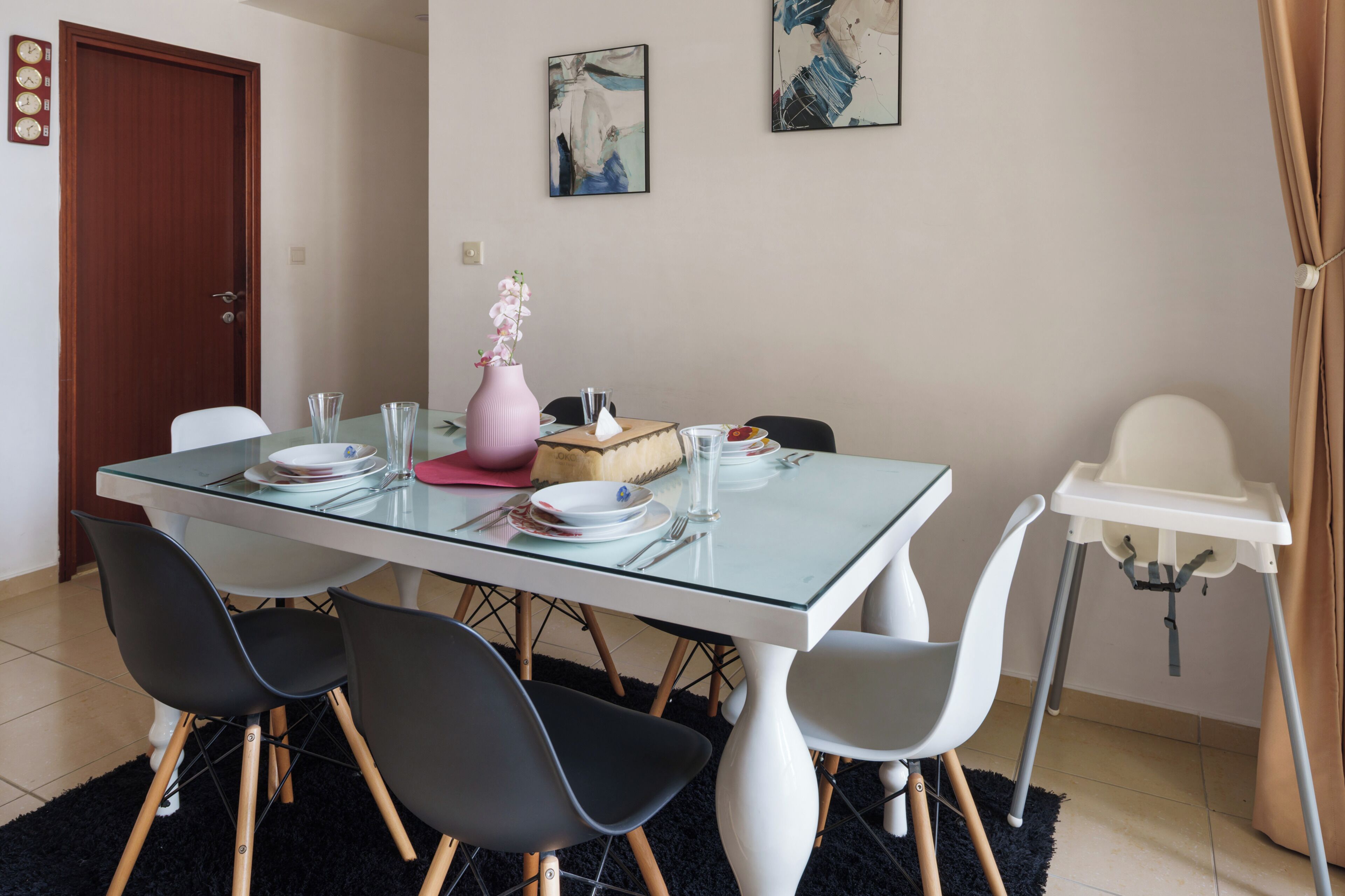 Un comedor con una mesa de vidrio, sillas blancas y negras, un florero rosa y una silla blanca para un niño.