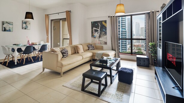 A modern living room with a sofa, coffee table, and TV, a dining area with chairs, and a large window with a city view. - Spacious 2 Bedroom Apt in Sadaf JBR  (Dubai)