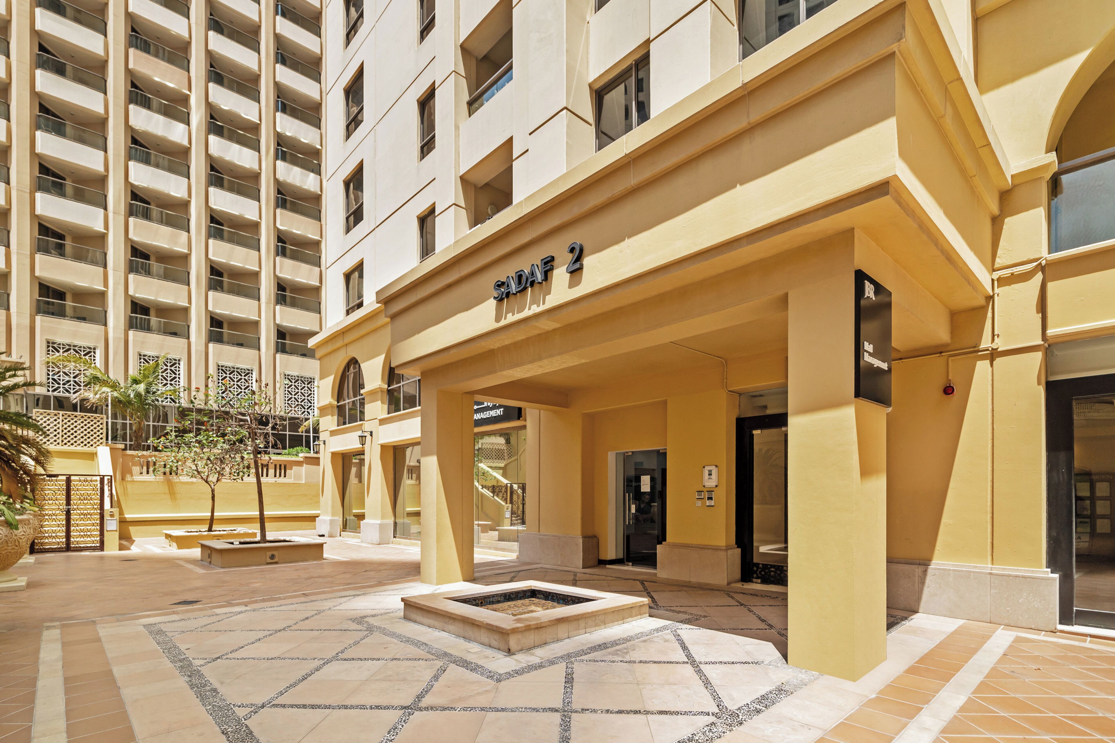 Entrada de un hotel con un letrero que dice 'SADAF 2', un espacio al aire libre cubierto y una fuente.