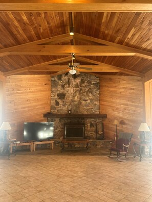 Une pièce dotée d’une cheminée en pierre, de poutres apparentes en bois au plafond, d’un ventilateur de plafond et d’un téléviseur posé sur un meuble en bois.