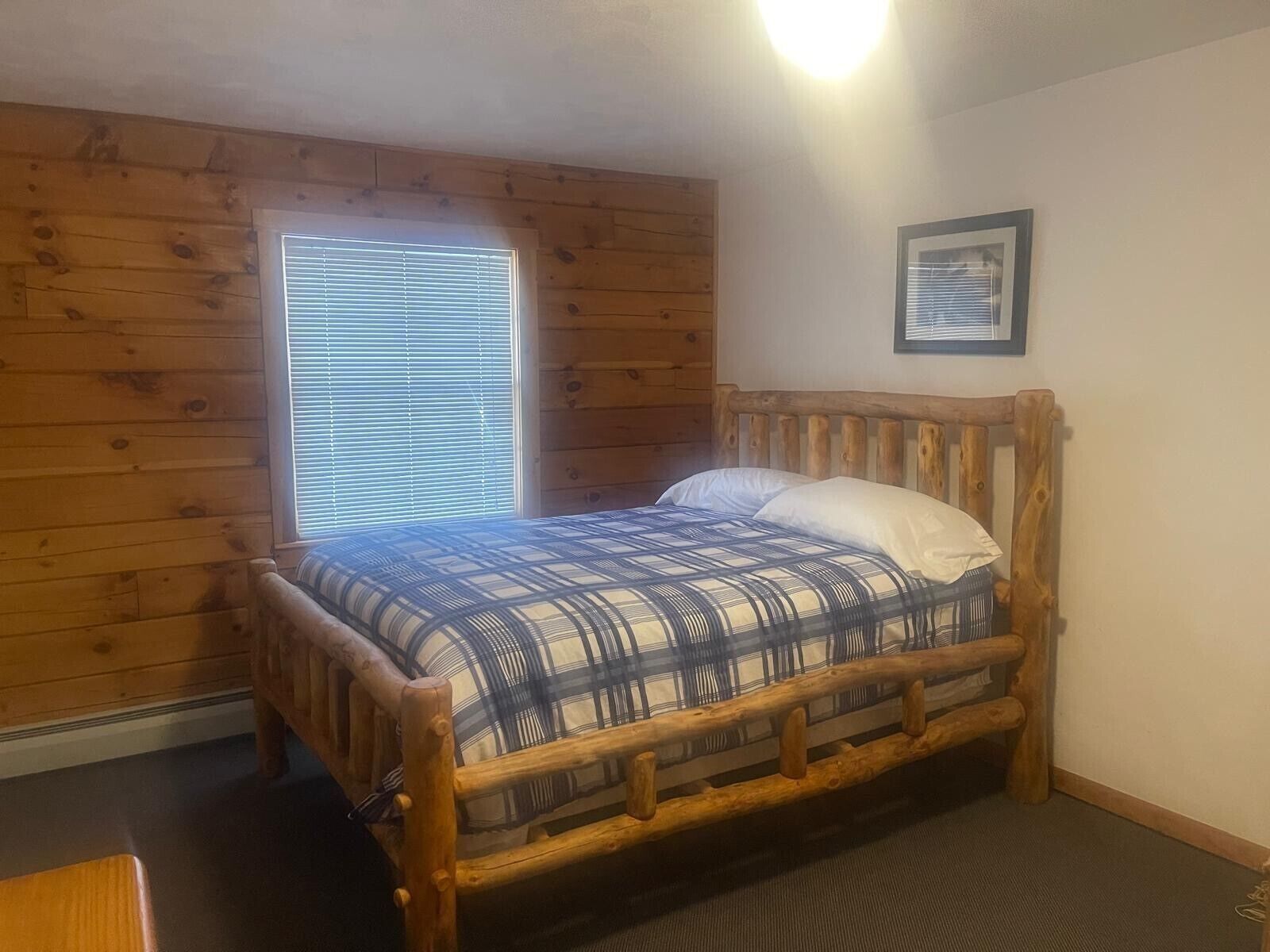 Una cama con estructura de madera y colcha de cuadros, una ventana con persianas, y un cuadro en la pared.