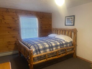 A wooden-framed bed with a plaid bedspread, a window with blinds, and a framed picture on the wall.